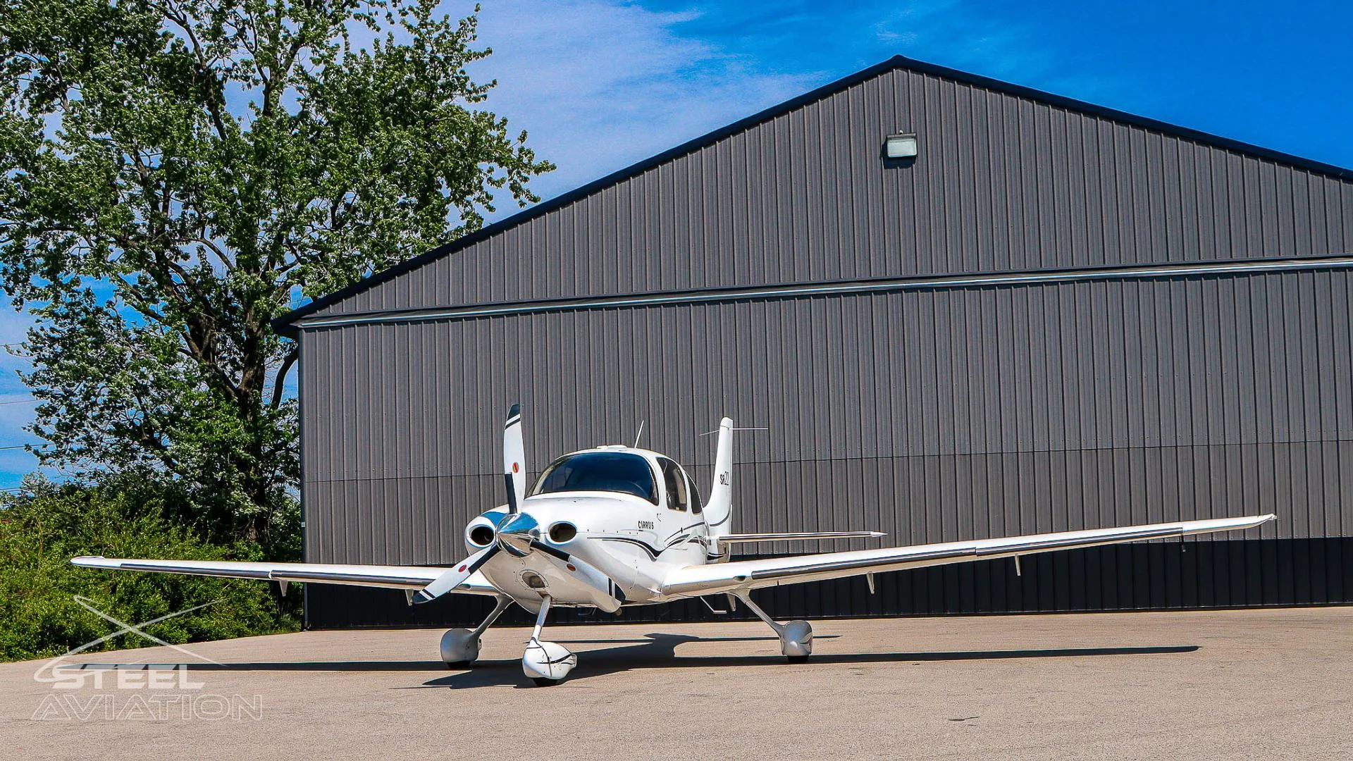 Front three-quarter view of a white Cirrus SR22 aircraft with black and grey stripes, parked in front of a grey hangar.