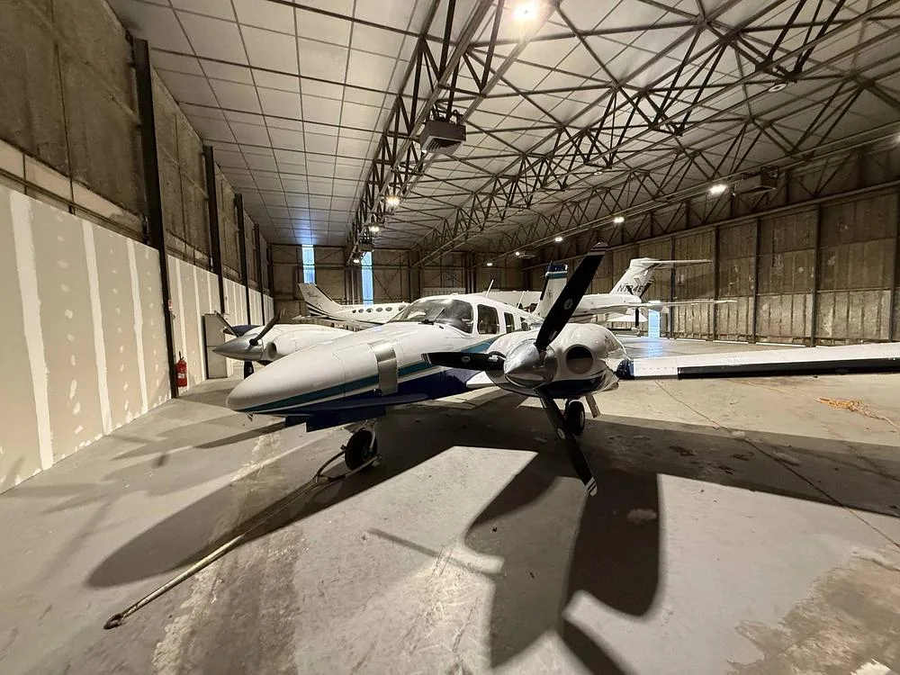 Front-left view of a white and blue Piper Seneca twin-engine propeller aircraft in a hangar.