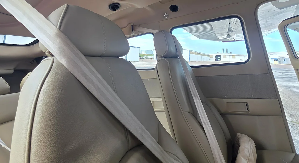 The tan leather interior of an aircraft cabin, showing two front seats with seatbelts and a view out the window.