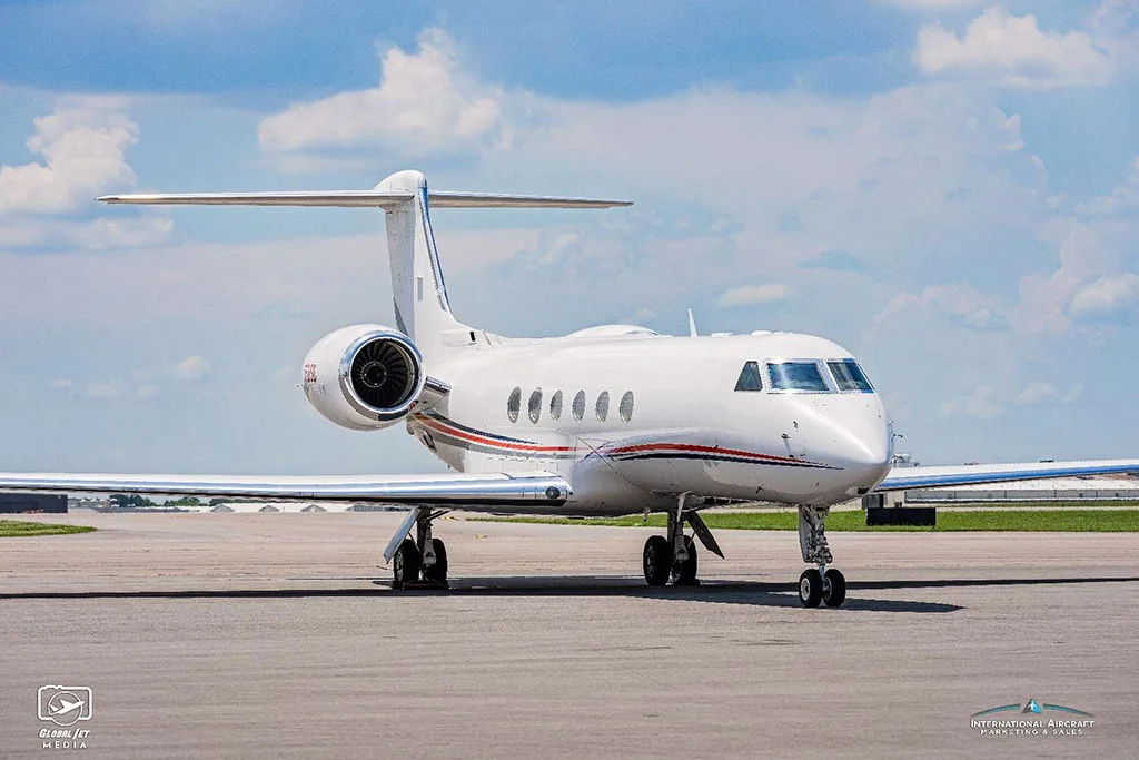 A front-quarter view of a white Gulfstream G550 aircraft with red and blue stripes, parked on an airfield.