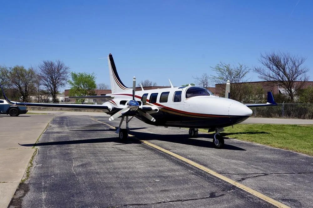 A front-quarter view of a white and dark blue twin-engine propeller aircraft with gold and red stripes, parked on an asphalt ramp.