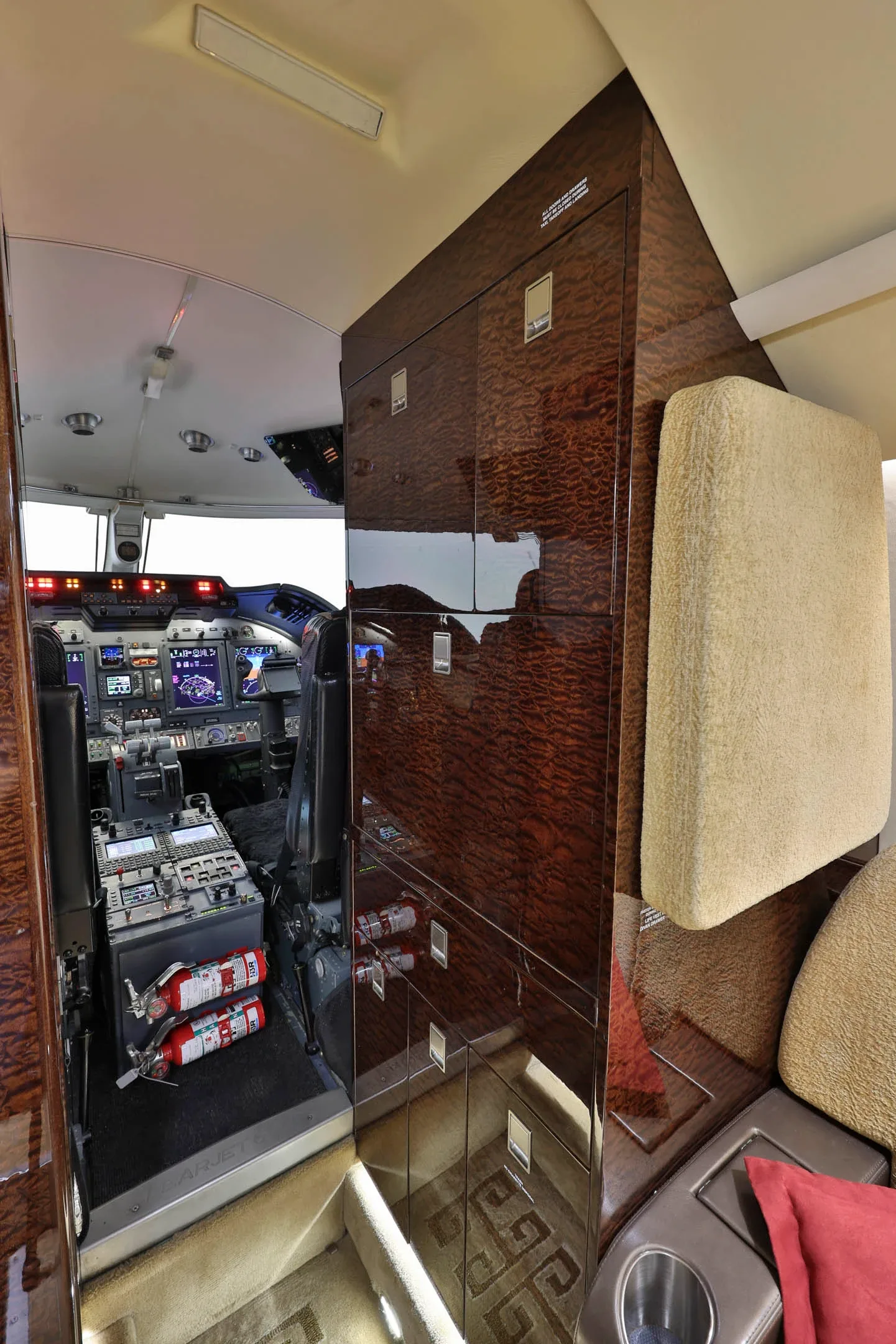 A luxurious aircraft cabin interior featuring a polished wood cabinet and a beige upholstered seat, with the cockpit visible beyond.