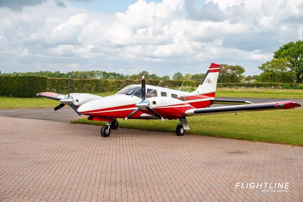 A red and white twin-engine Piper Seneca aircraft parked on a paved surface with grass and trees in the background.