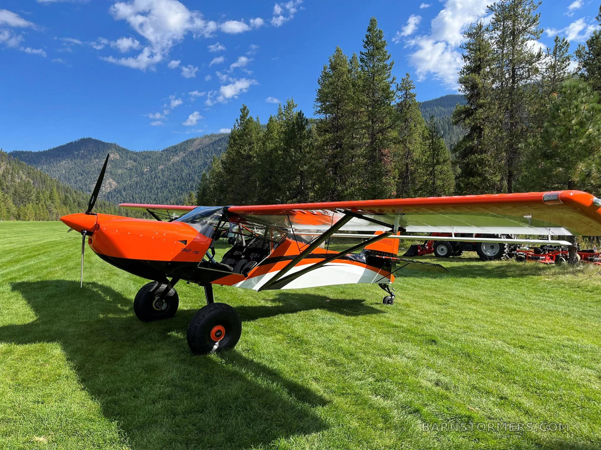 An orange and white Kitfox S7 Super Sport aircraft with large tires parked on a green grassy field with mountains and trees in the background.