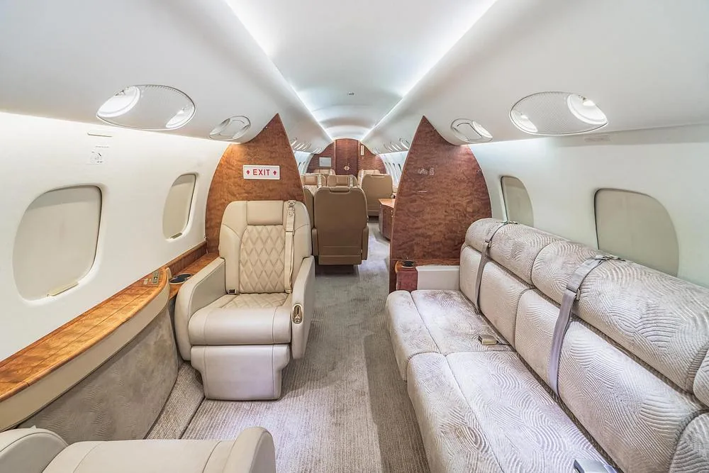 A view of the business jet cabin interior shows a beige couch and individual tan leather seats with wood paneling.