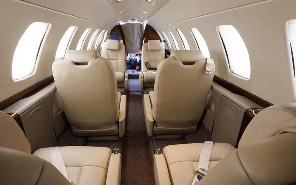 The interior cabin of a private jet with light tan leather seats, viewed from the rear looking forward.