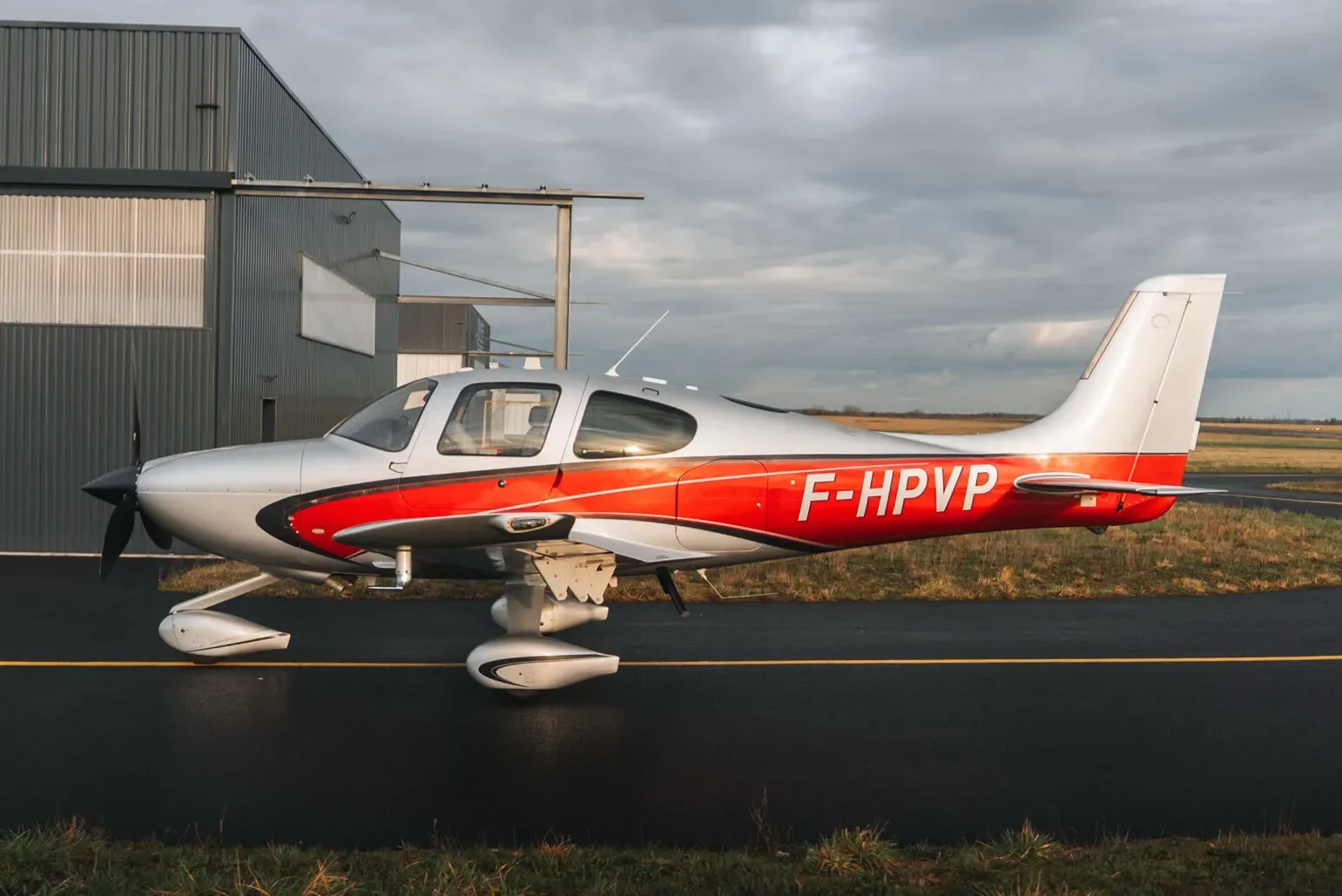 A silver and red Cirrus SR22 aircraft parked on an airfield next to a hangar under a cloudy sky.