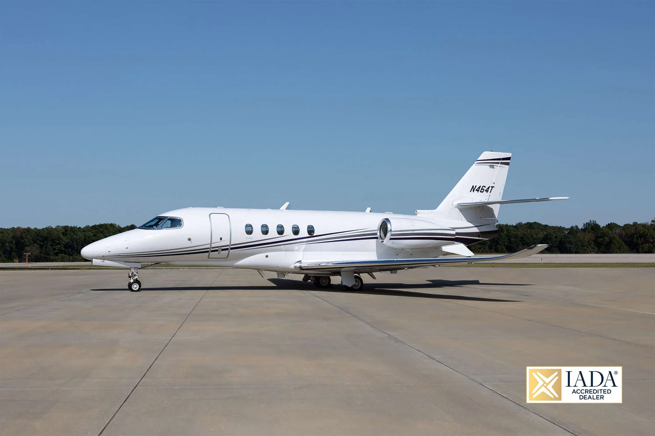 A white business jet with dark stripes, registration N464T, parked on an airfield under a clear blue sky.
