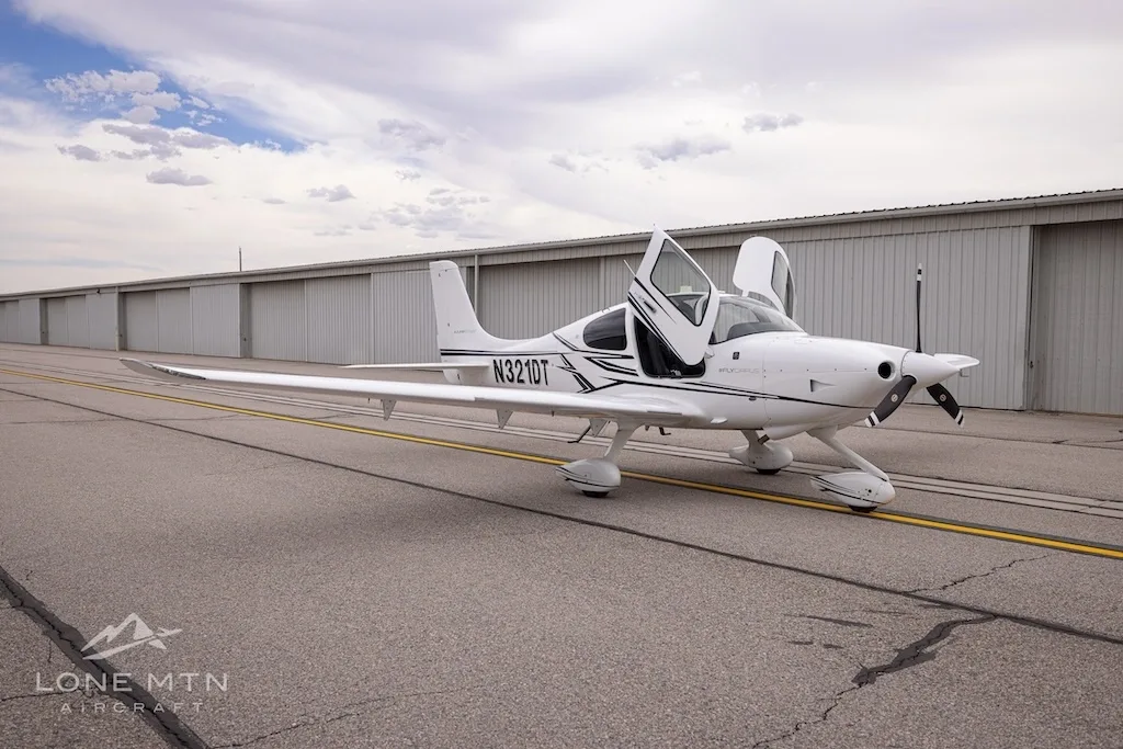 White Cirrus SR22 aircraft with black and grey stripes, doors open, parked on an asphalt ramp with hangars in the background.