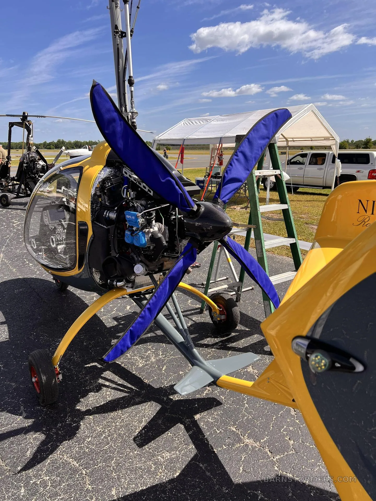 A yellow and grey gyrocopter with blue propeller covers, parked on an asphalt runway with tents and other aircraft in the background.
