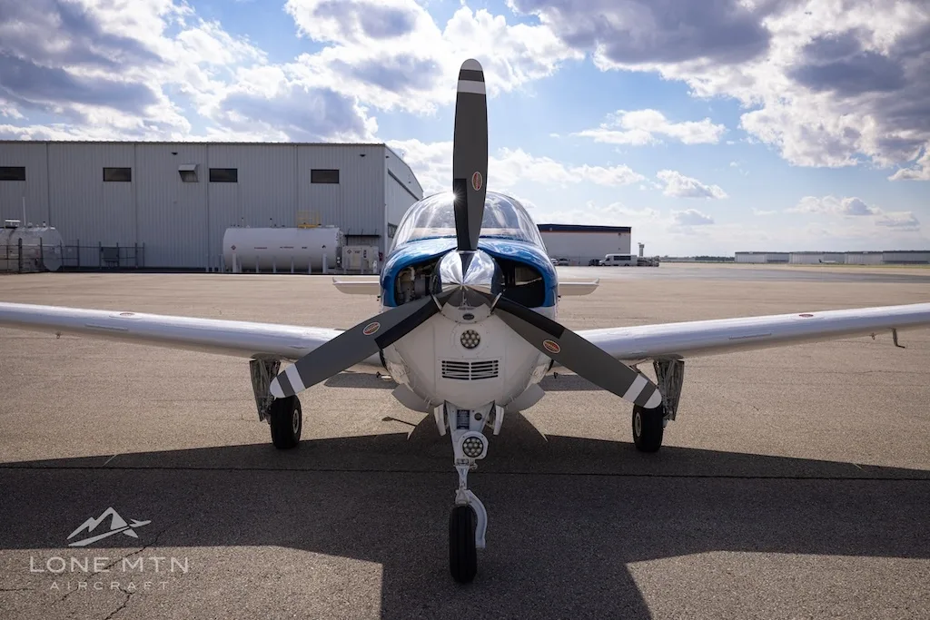 A head-on view of a white, blue, and red Beechcraft Bonanza N680JP with its propeller in front of a hangar.
