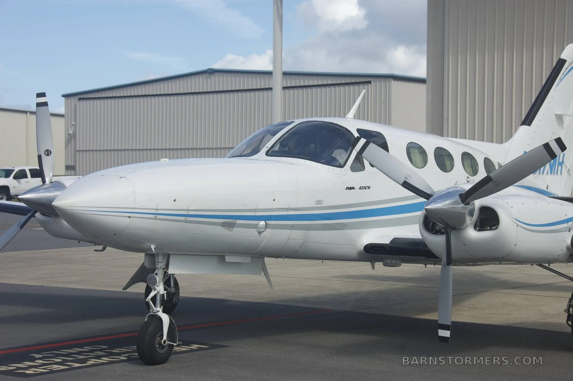 A close-up front-right view of a white twin-engine propeller aircraft with blue and grey stripes (N517MH) parked on a tarmac.