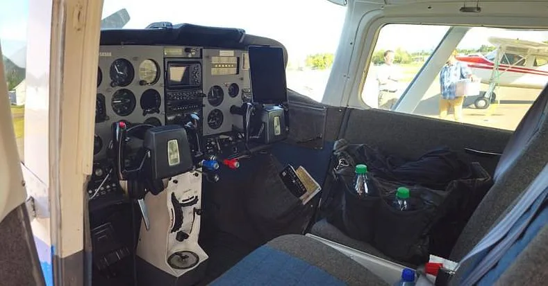The interior of the aircraft, showing the cockpit panel, pilot's seat, and a bag with water bottles on the passenger seat.