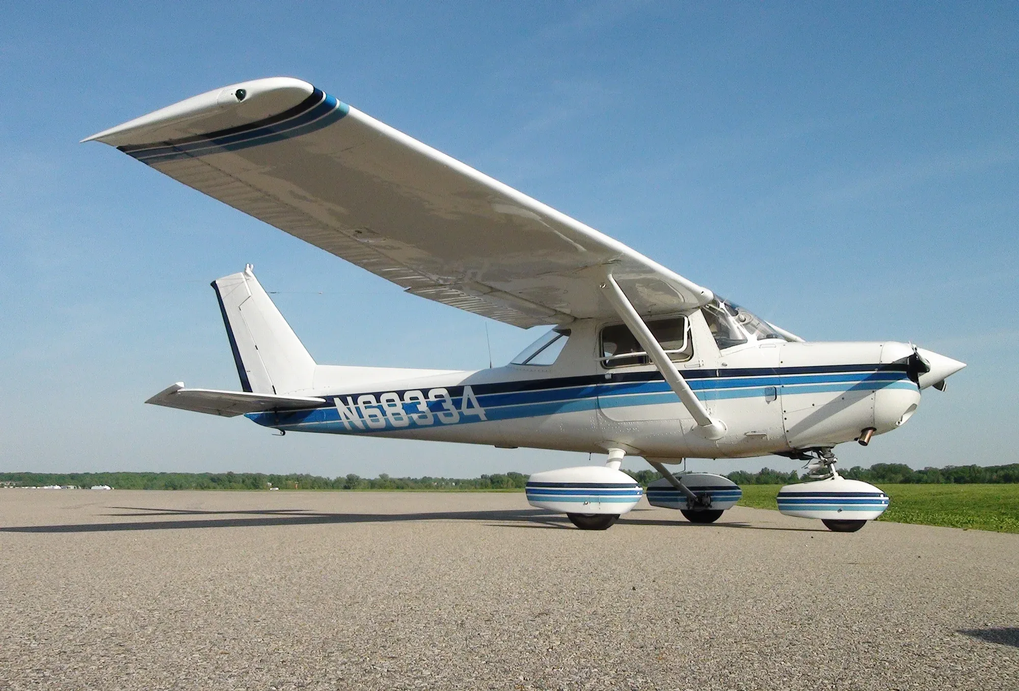 A white single-engine high-wing aircraft with blue stripes (N68334) parked on an asphalt surface under a clear blue sky.