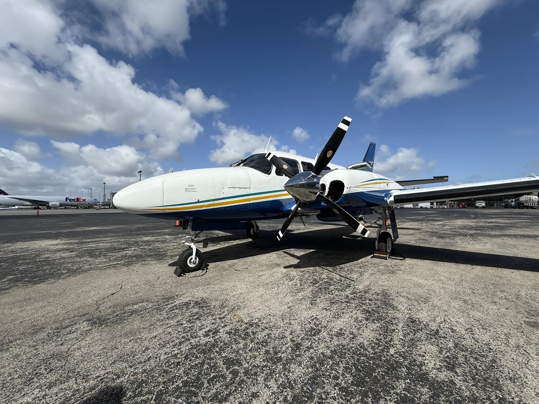 A white and blue twin-engine propeller aircraft with yellow and green stripes on the fuselage, parked on an airport tarmac under a partly cloudy sky.