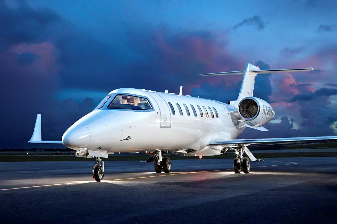 A white business jet with a dark blue tail stripe, parked on a tarmac at dusk with dramatic clouds.