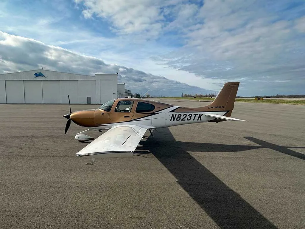 A Cirrus SR22 aircraft, N823TK, with a brown and white paint scheme parked on an airfield next to a hangar.