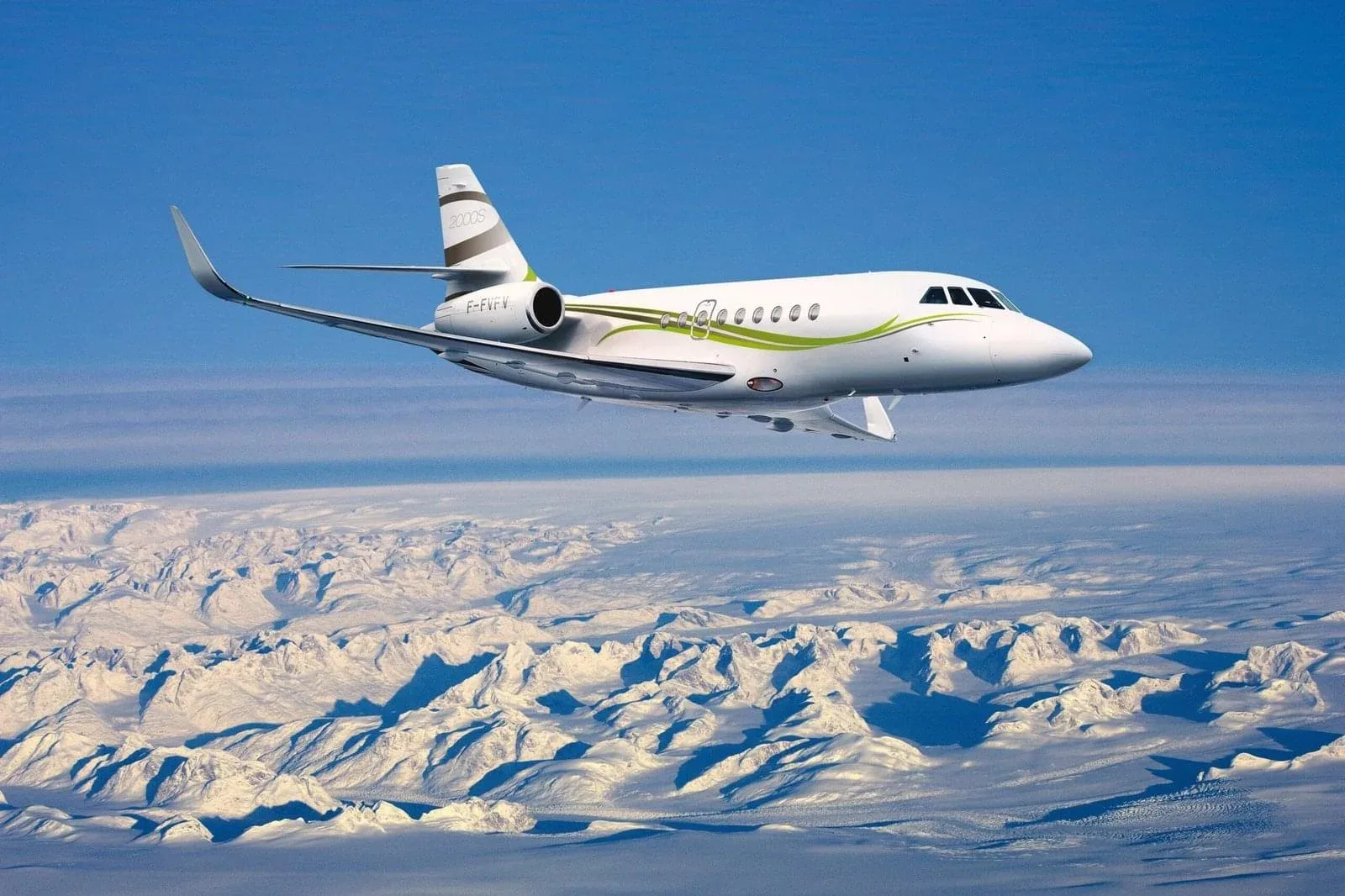 A white Dassault Falcon 2000S business jet with green and brown stripes flies over snow-covered mountains under a clear blue sky.
