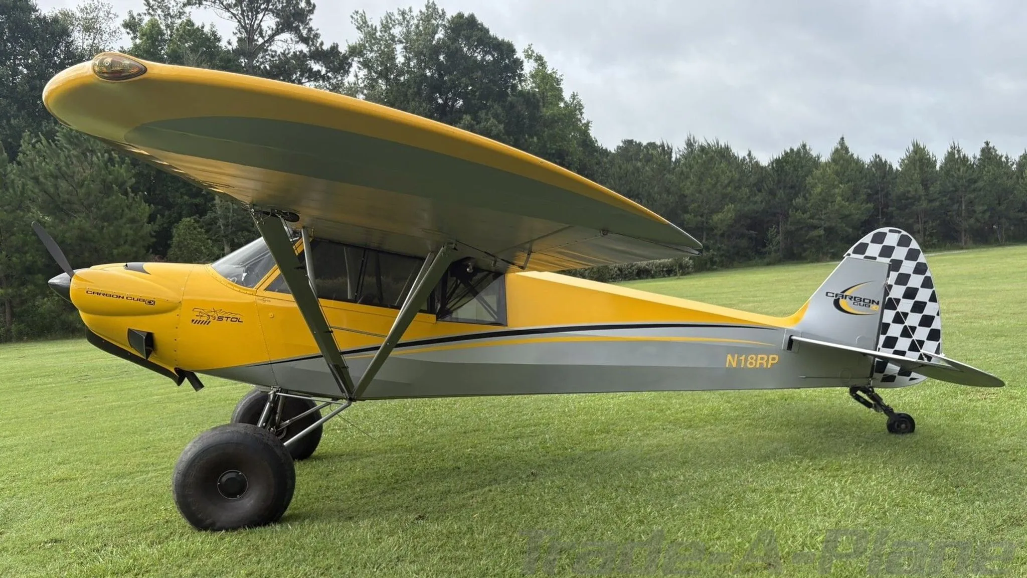 A side view of the yellow and gray CubCrafters Carbon Cub aircraft N18RP, showing its checkered tail and large tundra tires, parked on grass.