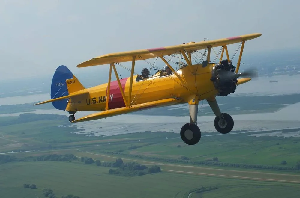 A yellow Stearman biplane with red and blue accents, marked 'U.S. NAVY' and N1344V, flying over a body of water and green landscape.