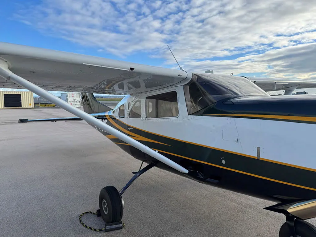A close-up view of the front fuselage and wing of the dark green and white Cessna 182, showing the propeller and landing gear.