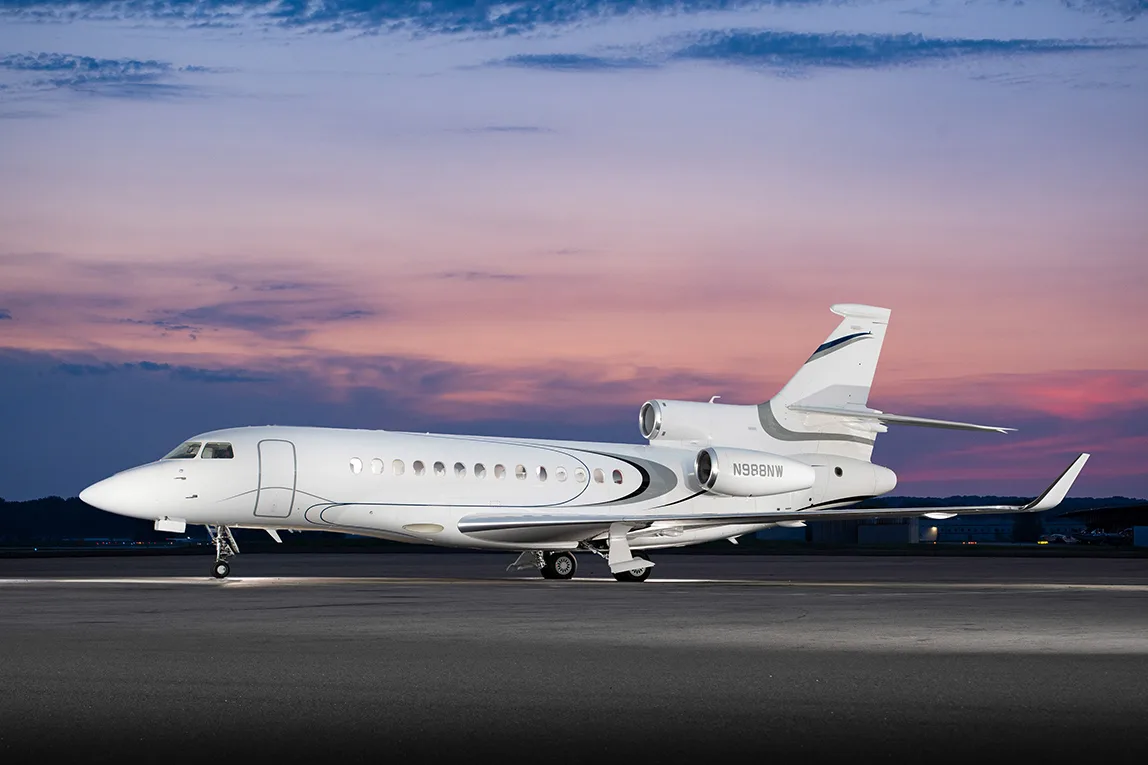 A white Dassault Falcon 900EX business jet with grey and black stripes parked on a tarmac at dusk.