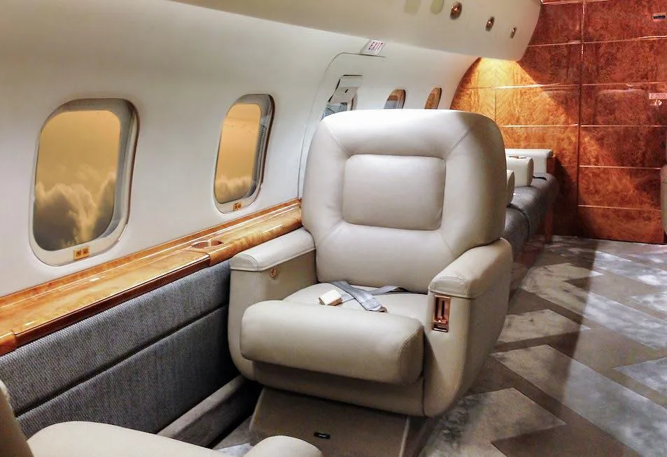A beige leather seat in a private jet cabin, with wood paneling and windows showing clouds outside.