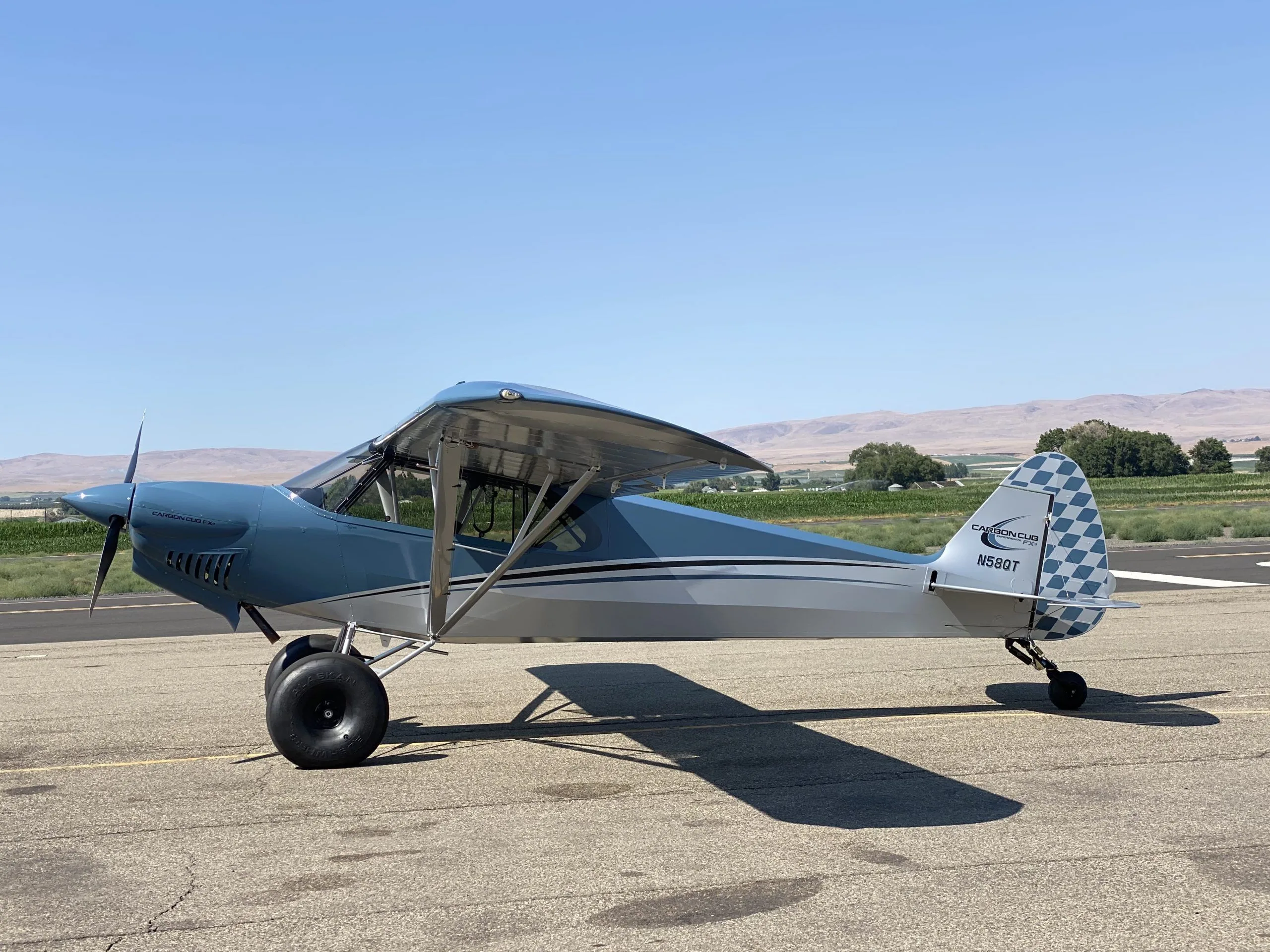 A blue and grey Carbon Cub FX-3 aircraft with registration N58QT parked on a tarmac with a rural landscape in the background.