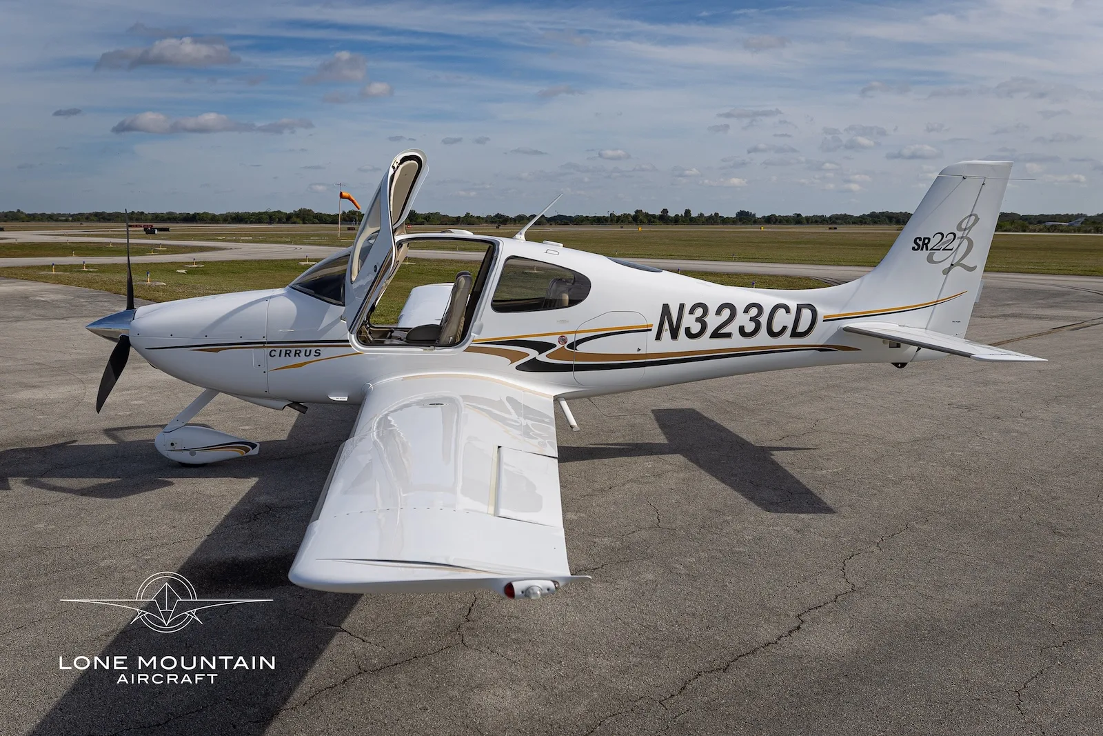 A white Cirrus SR22 aircraft with gold and black accents, registration N323CD, parked on an airfield with its doors open, viewed from the rear right.
