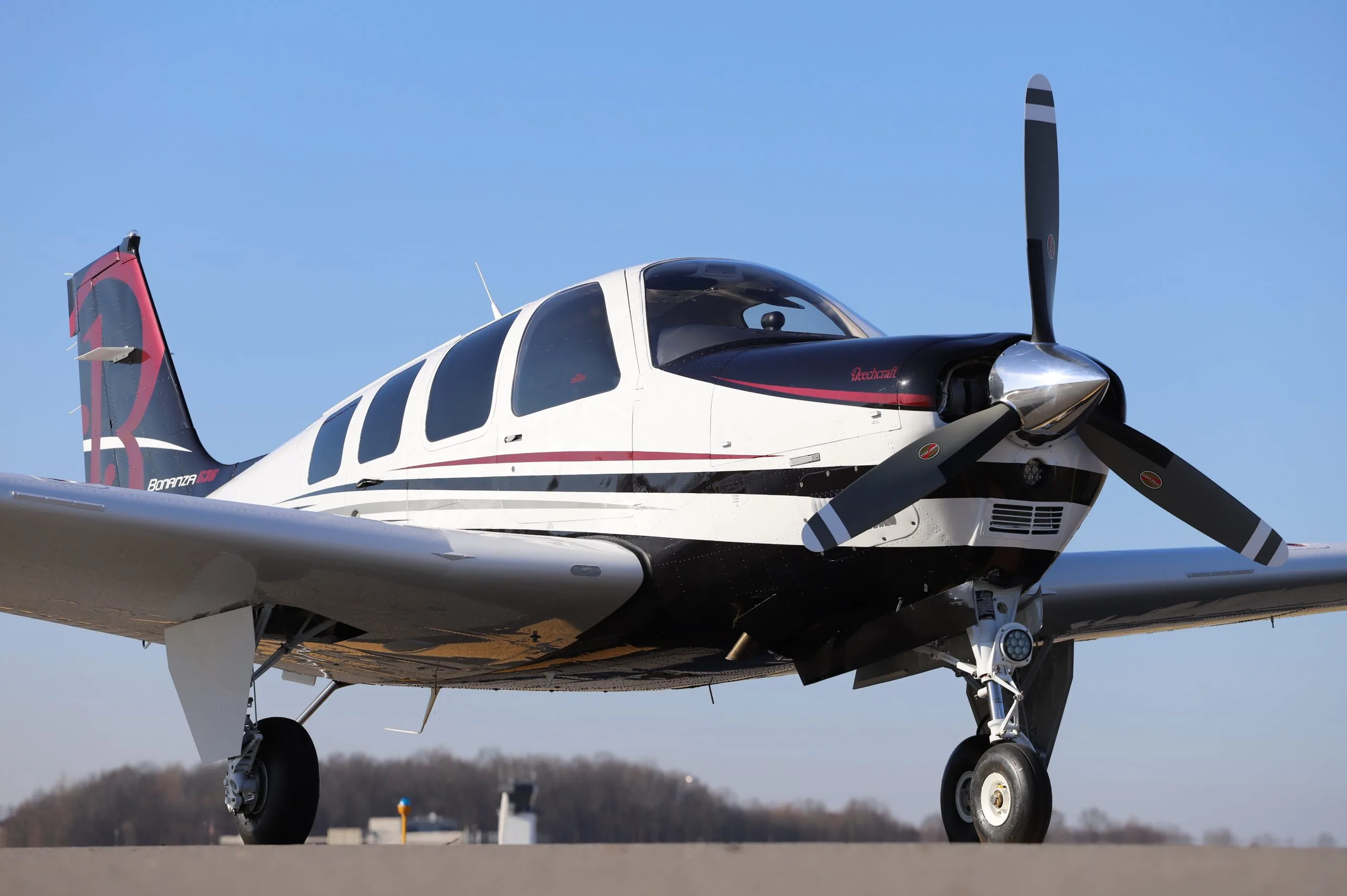 A white, black, and red Beechcraft Bonanza G36 with a large 'R' on its tail, parked on a tarmac under a clear blue sky.