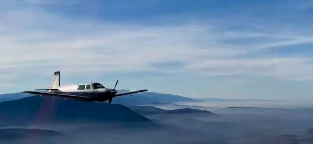 A blue and white single-engine aircraft flying over misty mountains under a blue sky.