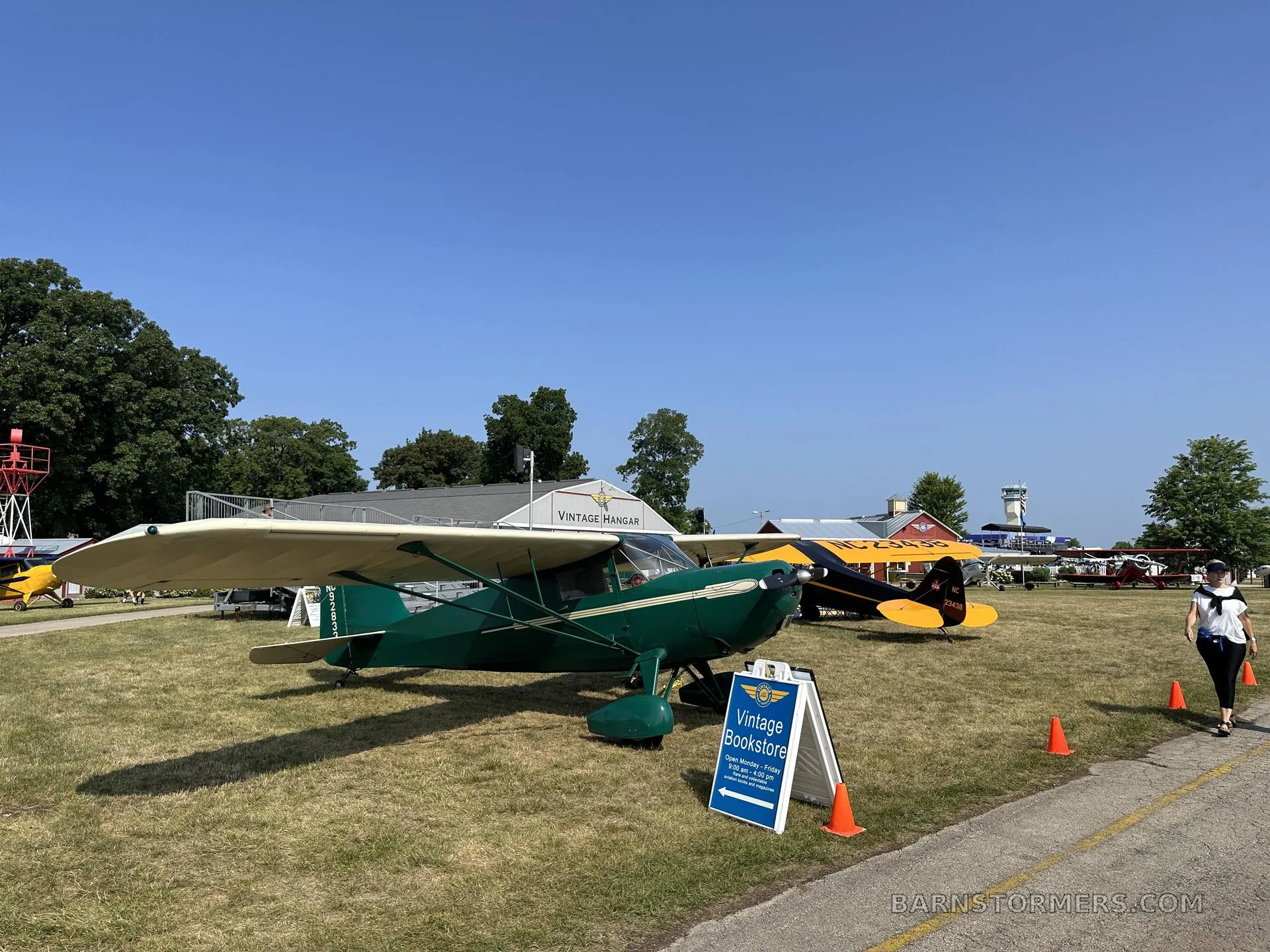 A green and cream vintage aircraft with tail number NC92833 is parked on grass at an airfield, with other aircraft and buildings in the background.