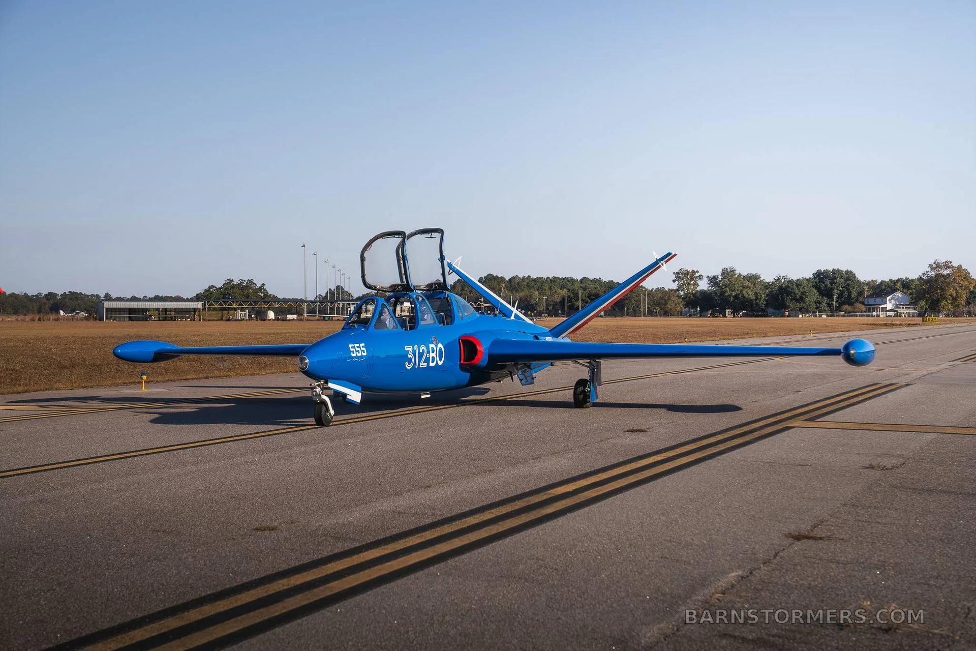 A blue Fouga CM.170 Magister jet aircraft with open canopies is parked on an asphalt runway, viewed from a slightly different angle.