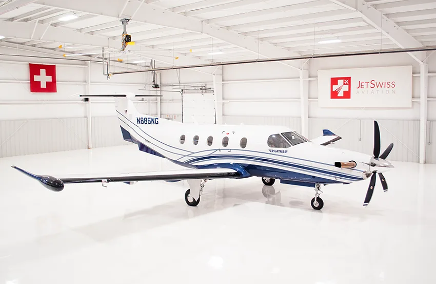 A white Pilatus PC-12 with blue and silver stripes is parked inside a brightly lit white hangar.