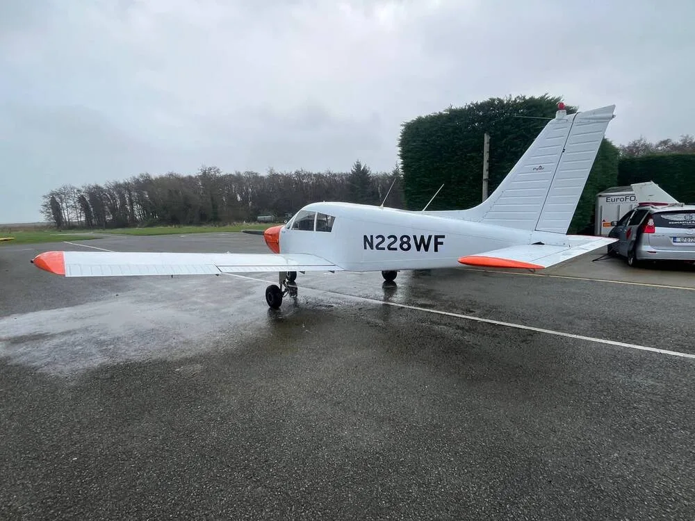 A white and orange Piper Cherokee 140 (N228WF) parked on a wet tarmac at an airfield, viewed from the rear-left.