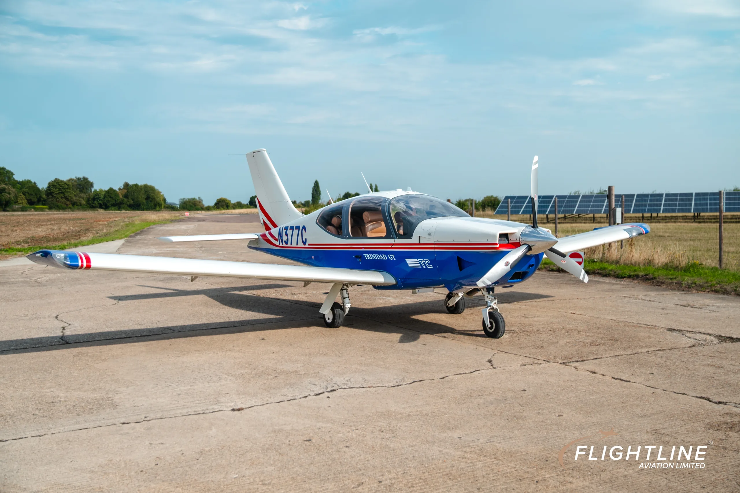 A front-right view of the N377C Socata Trinidad GT, painted white and blue with red stripes, parked on a concrete pad with solar panels in the background.