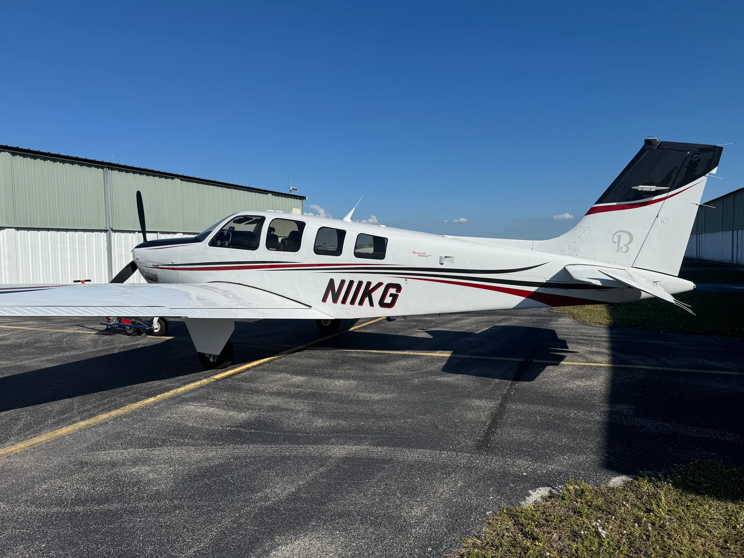 A white Beechcraft Bonanza N11KG with black and red stripes parked on an asphalt ramp under a clear blue sky.