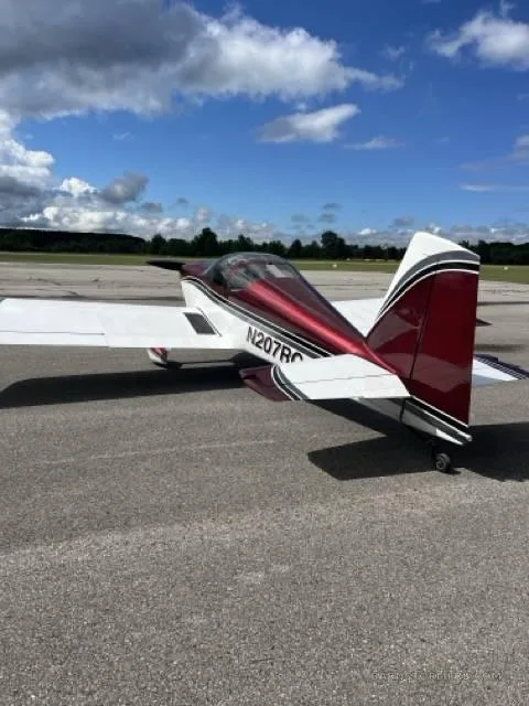A rear-side view of a red and white single-engine aircraft with tail number N207RC, parked on an asphalt runway under a blue sky.