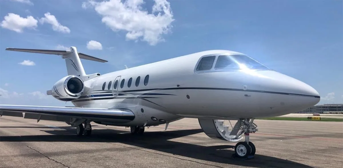 A white business jet with dark blue and grey stripes on the tarmac under a partly cloudy sky.