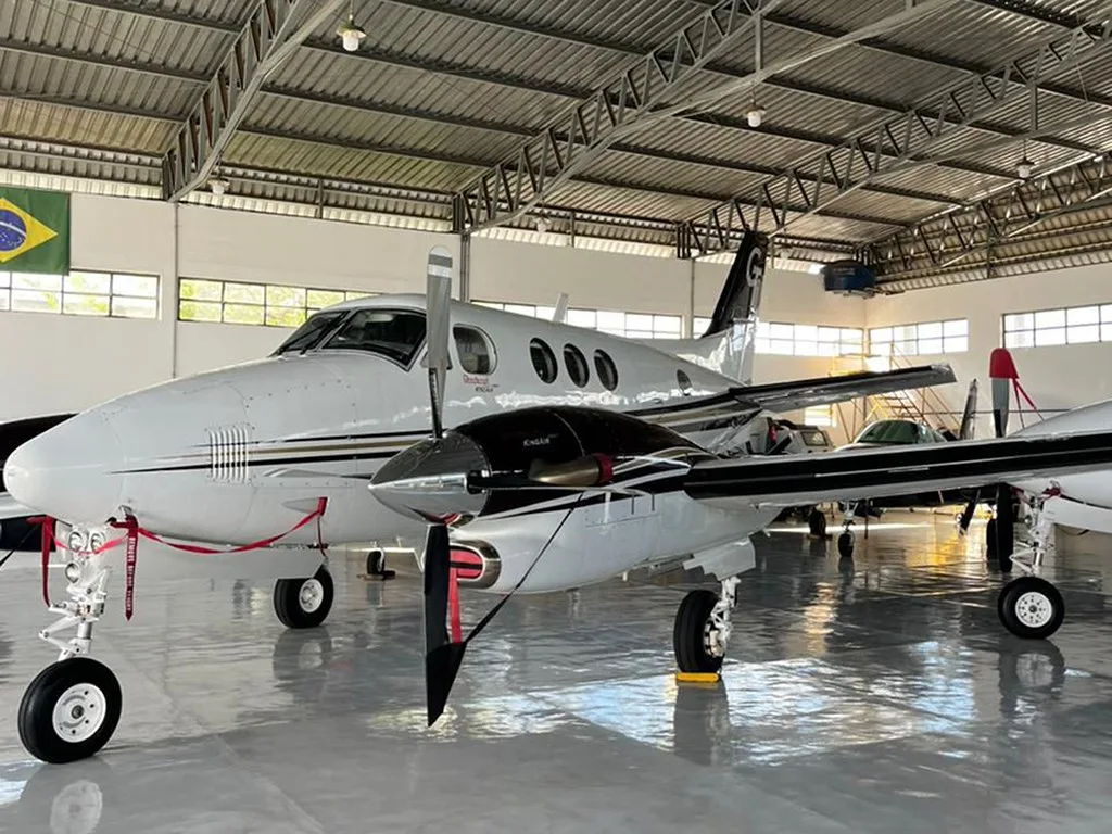 A white and black Beechcraft King Air 350 with gold stripes, seen from the front-left in a hangar, with a Brazilian flag in the background.