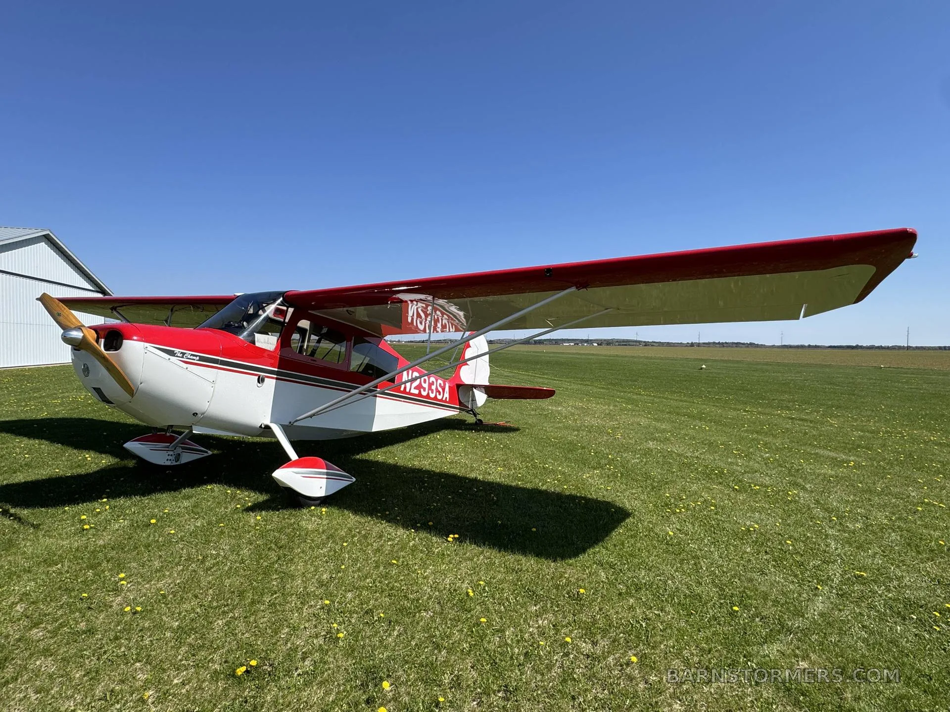 A red and white single-engine aircraft with tail number N293SA parked on a grassy field next to a hangar.