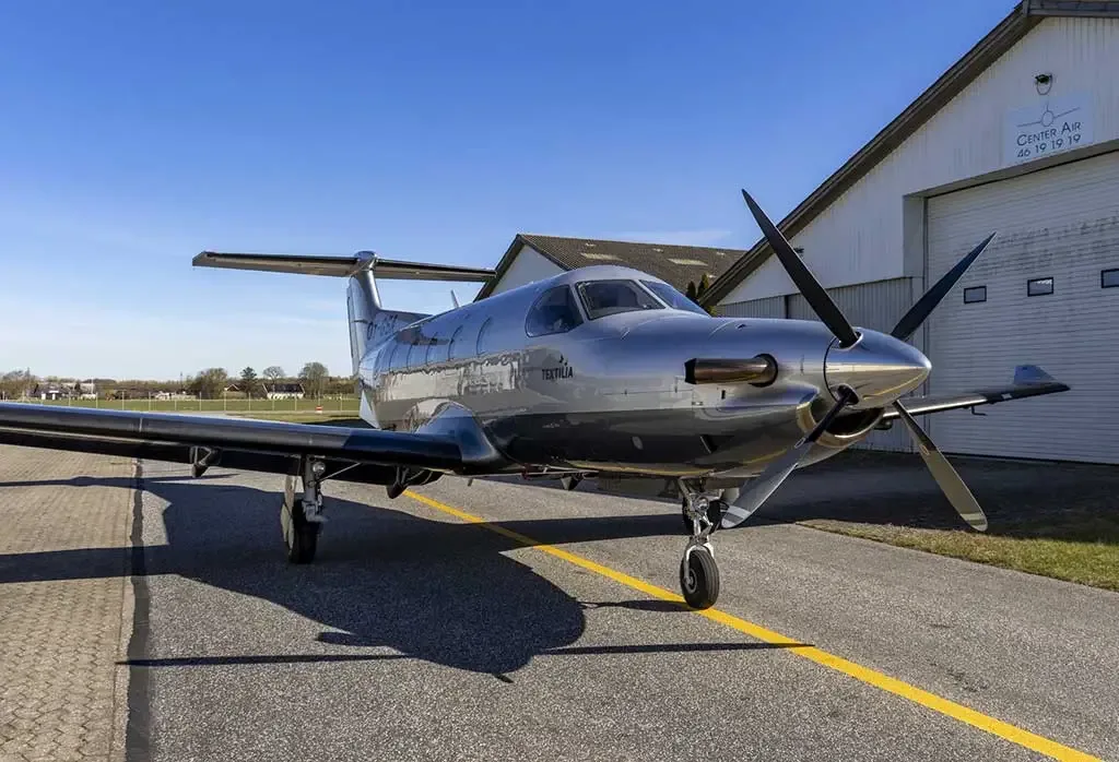 A silver and dark grey Pilatus PC-12 aircraft with a four-bladed propeller, parked on an asphalt tarmac in front of a hangar.