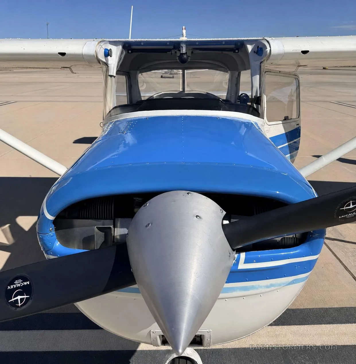 A head-on view of a white and blue single-engine propeller aircraft, showing the propeller and front cowling, parked on an airfield.