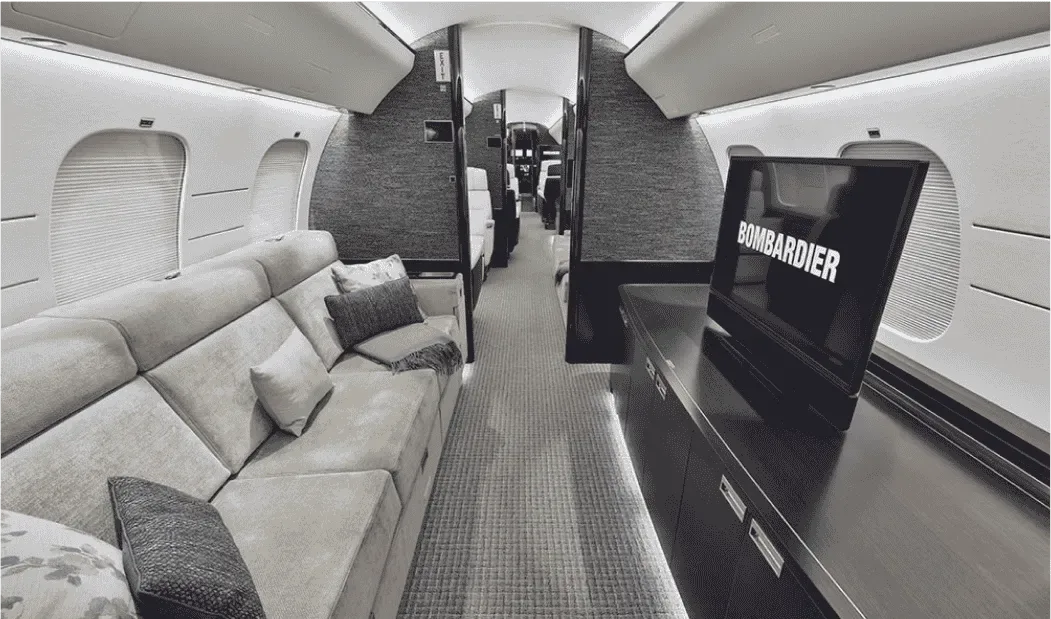 A luxurious aircraft cabin interior with a large sofa on the left and a TV displaying "BOMBARDIER" on the right.