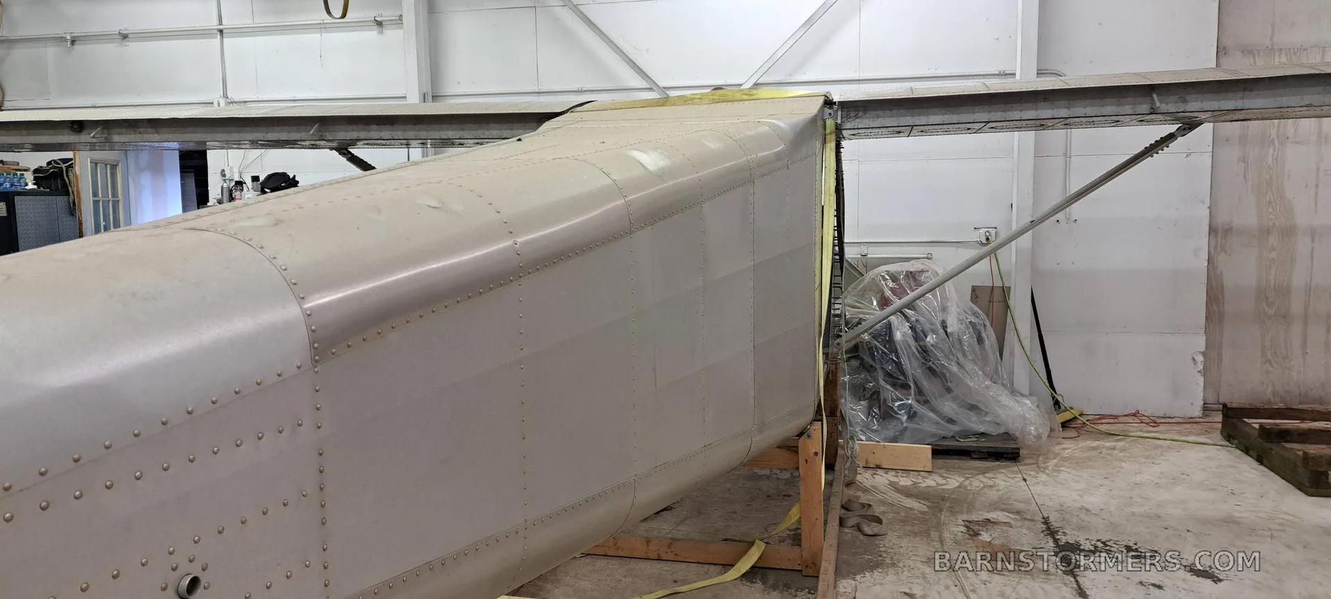 A partially assembled aircraft fuselage and wing, made of bare metal, supported on wooden stands in a workshop.