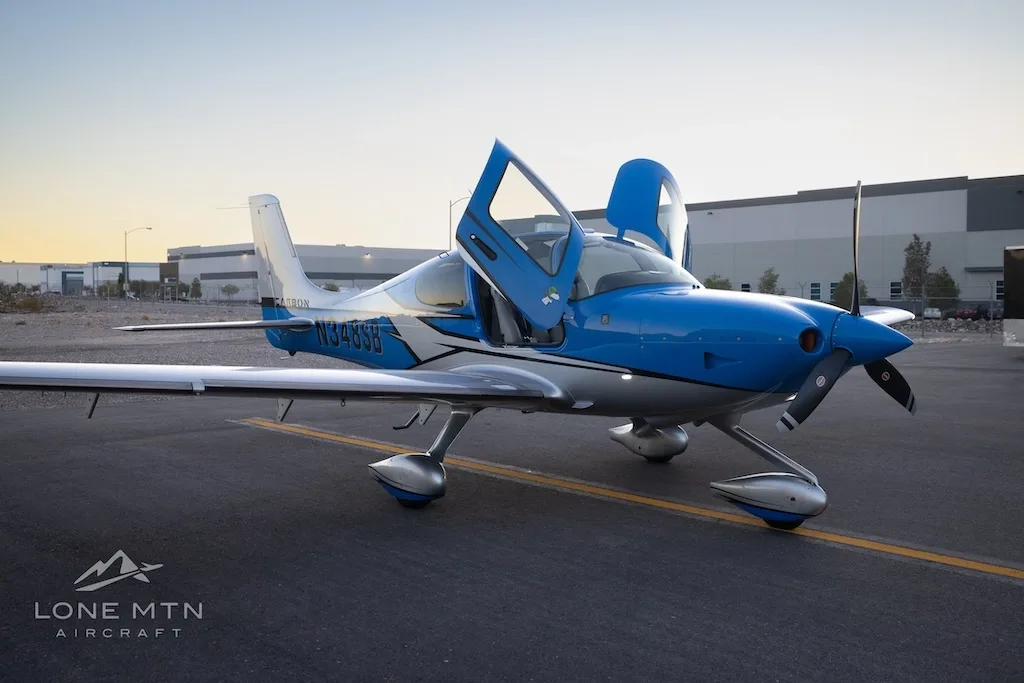 A blue and silver Cirrus SR22 aircraft with open doors parked on an airfield with hangars in the background at sunset.