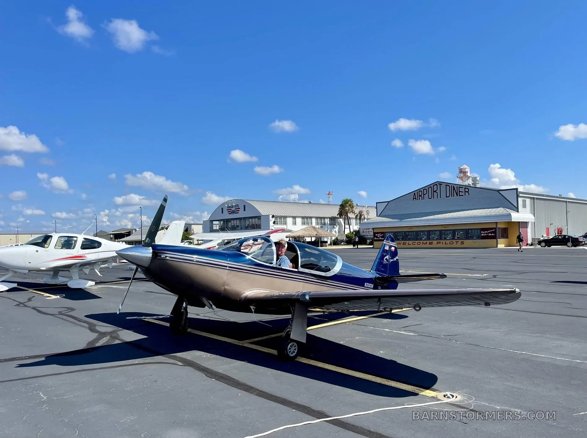 A bronze and dark blue single-engine aircraft with a pilot in the cockpit, parked on an airport tarmac next to an "Airport Diner" building.
