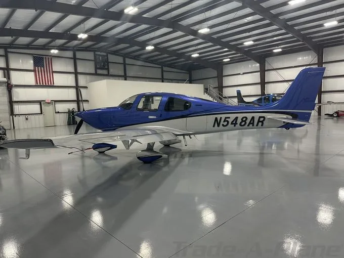A blue and white single-engine aircraft, N548AR, is parked inside a brightly lit hangar with an American flag on the wall.