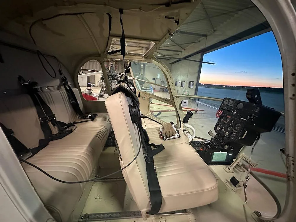 The interior of the Hiller UH-12 helicopter with beige seats and instrument panel, looking out of a hangar at dusk.