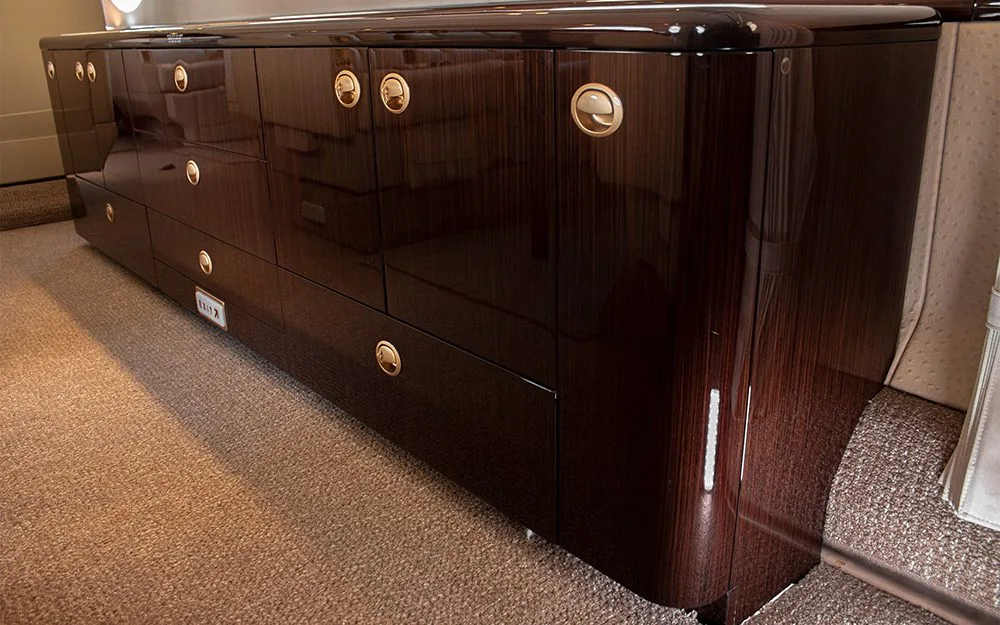 A long, dark wood credenza with gold hardware and multiple drawers and cabinets inside an aircraft cabin.